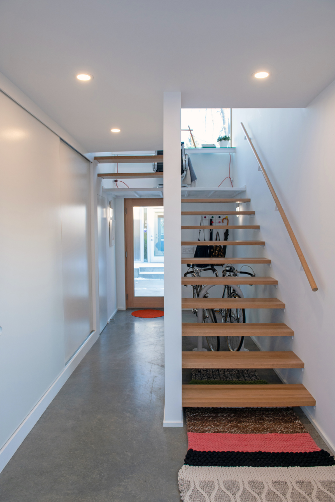 A slim entryway with concrete floors, white walls, sliding door storage closets, simple wooden floating stairs with a matching stair rail and plenty of natural light via large windows.