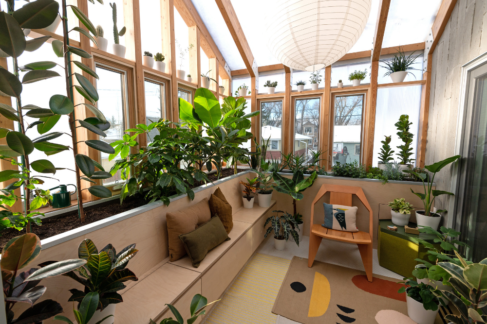 A glass and wood solarium with planters built in around the perimeter of the room housing countless lush green plants. The floor is layered with abstract rugs and built-in seating with storage as well as a couple chairs encircle the room.