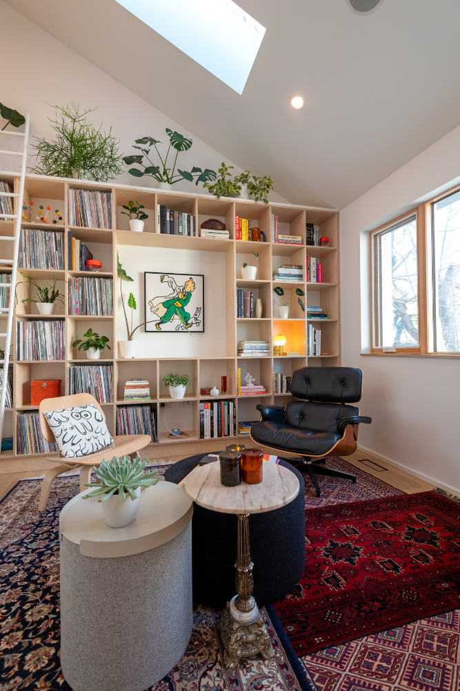 Living room with layered Persian rugs, several side tables clustered together, mismatching chairs and built-in bookshelves full of records, art, plants and knickkancks.