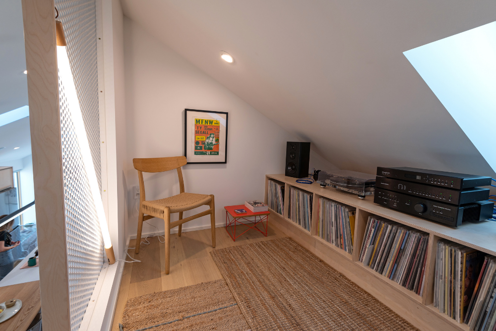 The interior of the loft, featuring jute rugs, a wood shelf full of records with a turntable and music player system sitting above it, a white mesh wall with a light bar mounted diagonally across it casting soft white light around the room, a wood and rattan chair, a small bright red table, a framed print and a skylight.