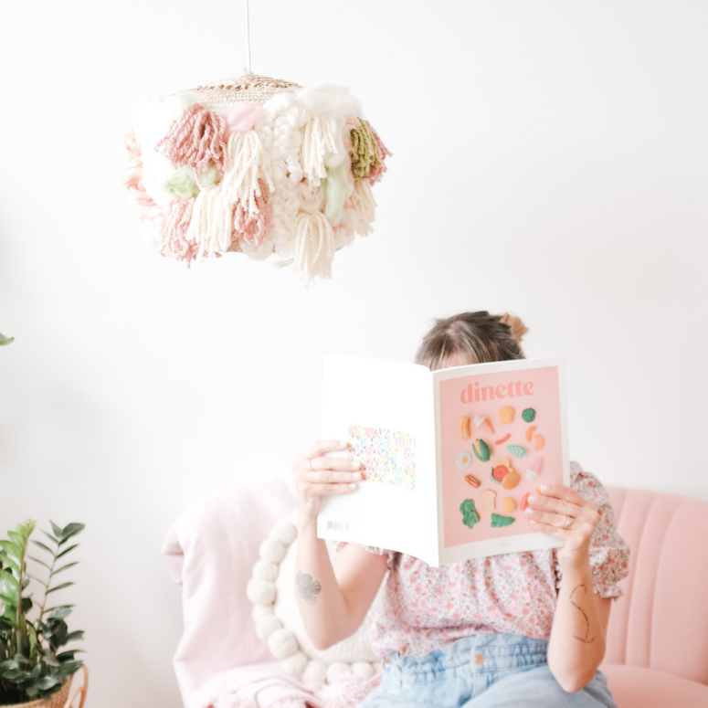 The DIY wicker basket lampshade covered in pink, green and white wool details hangs over the DIY creator, Maca, as she sits on a pink couch reading a Dinette magazine.