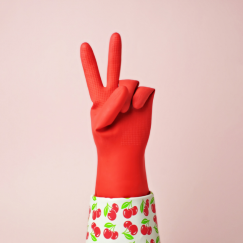 A hand in a red rubber cleaning glove with a cherry-print trim, throwing up the peace sign in front of a pale pink wall.
