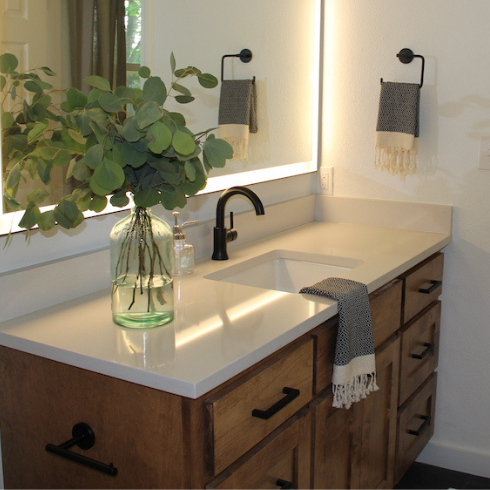 A clean, empty sink with clean white countertops clear of clutter, with just a glass vase of eucalyptus trimmings sitting on it. A large rectangular LED-framed bathroom mirror hangs above.