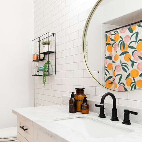 A white bathroom with toiletries in matching bottles on the sink, knickknacks on a small wall-hanging wire shelf above the toilet and a shower curtain printed all over with peaches, reflected in a round mirror hanging over the sink.