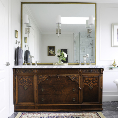An ornate vintage cabinet serves as the base for the sink in a grand bathroom. A huge gilded square-shaped mirror hangs above.