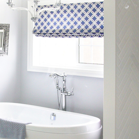 A bathroom window with blue and white patterned blinds is situated behind a white bathtub.
