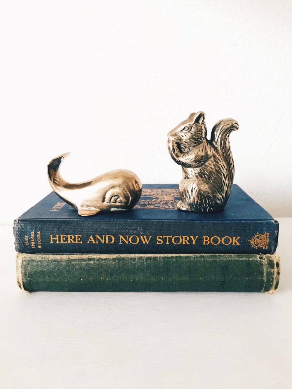 A pair of small brass figurines, a squirrel and a whale, sit atop a couple vintage books - one green with a worn spine and one navy blue with gold lettering on its spine.