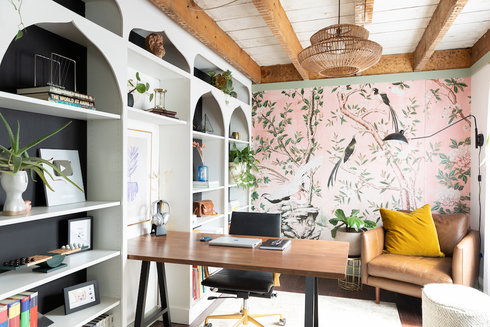 Designer Danie Berger's creative home office features a wall lined with built-in shelves and a bold pink wallpaper of birds and foliage on the back wall.