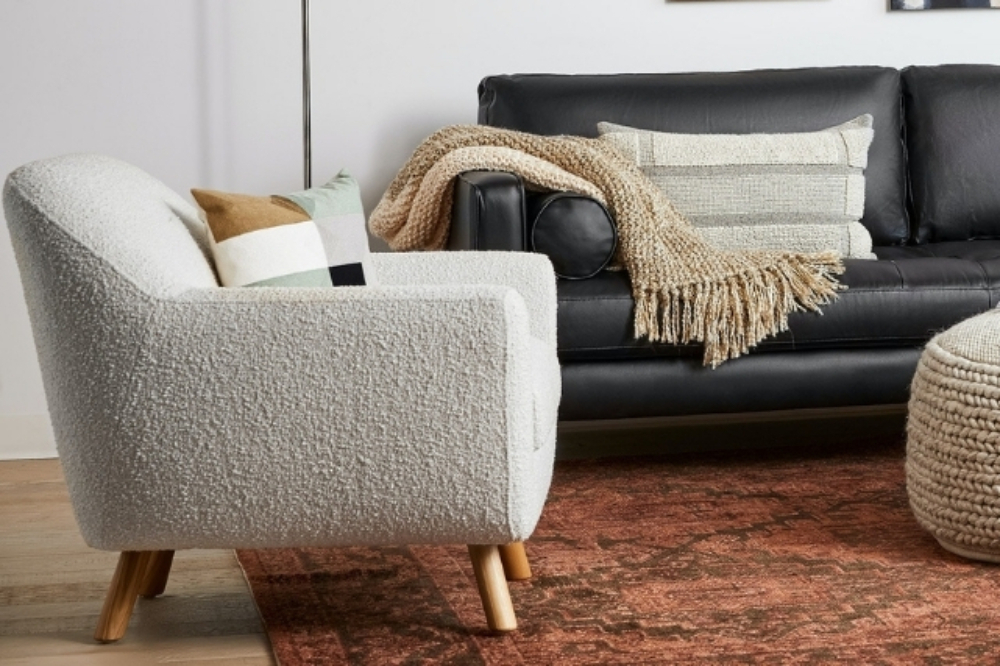 An ivory bouclé lounge chair with a colourful throw below and wooden legs, sitting beside a black leather sofa draped with a fluffy cushion and a throw blanket. Both seats, along with a chunky ottoman, sit on a rustic orange-ish Persian rug.