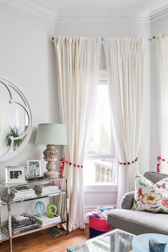 A cozy, eclectic living room rich in texture and colour receives lots of bright light from a window flanked by long white linen curtains tied back with garlands of colourful pompoms.