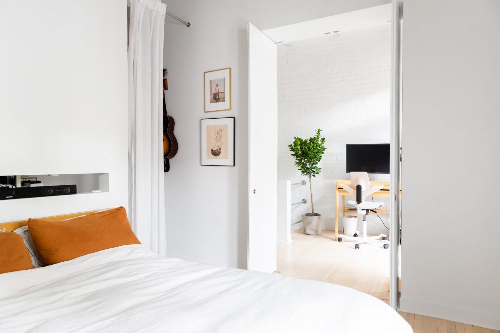 white bedroom with white bed and orange pillowcases, with view into office space in adjacent room