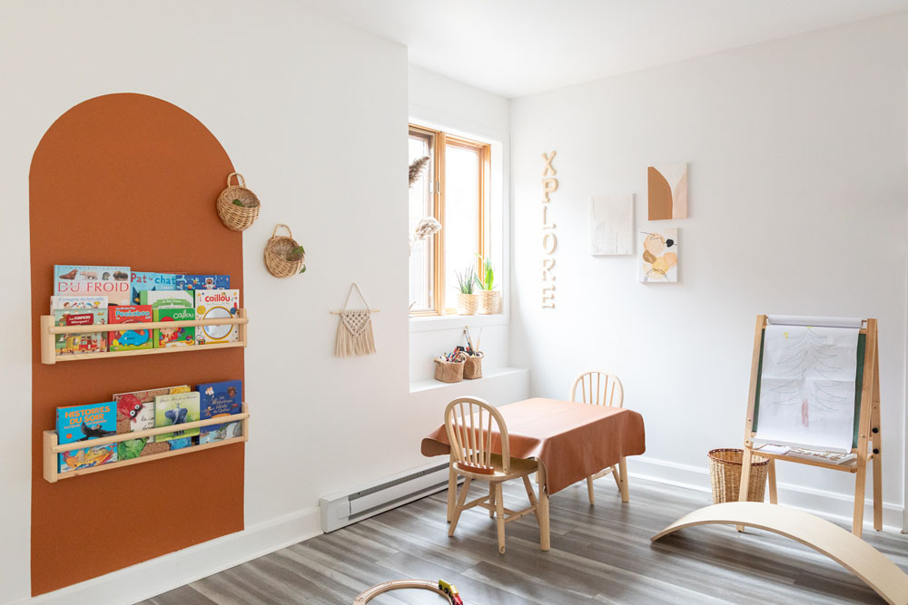A children's playroom with touches of orange, such as a painted orange arch on the wall behind some wall-mounted bookshelves full of children's books, an orange tablecloth over a little work table with two wooden chairs and orange-ish decor such as woven baskets, wooden toys and wall-hung paintings.