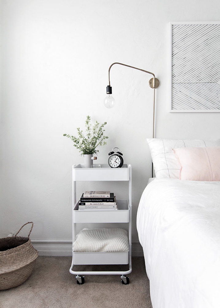 A white IKEA RASKOG utility cart is used in this bedroom as a three-tier nightstand, housing an extra blanket in the bottom tray, a stack of novels in the middle tray and an alarm clock and vase of fresh flowers in the top tray.