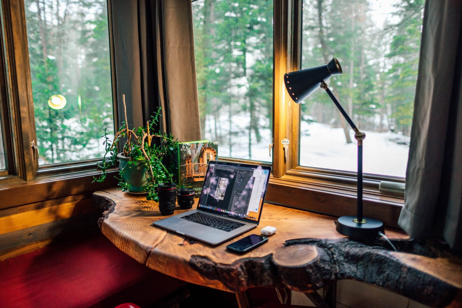 A live edge desk is pushed up against a bay window with a view of snow covered land and green fir trees. Atop the desk is a laptop, a desk lamp, and some small indoor plants.