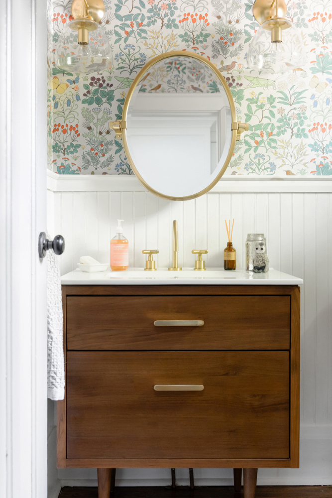 A powder room that boasts vibrant floral print wallpaper, plus a wooden vanity, white fixtures and brushed gold hardware to ground the space with timeless neutrals.