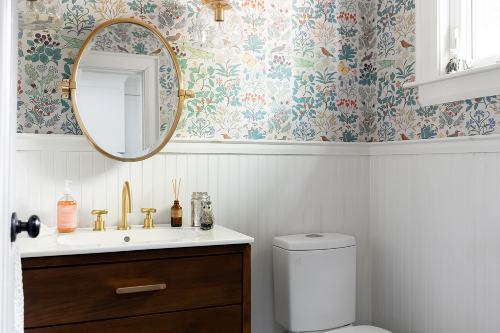 A fresh powdery room with white beadboard, a dark wooden vanity, brushed gold hardware and a colourful wallpaper featuring foliage, birds and insects like butterflies and grasshoppers.