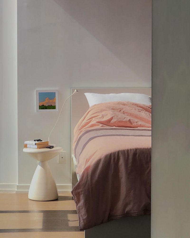 A small, sculptural nightstand resembling an hourglass shape holds several books and a lamp next to a bed with pink-tone bedding.