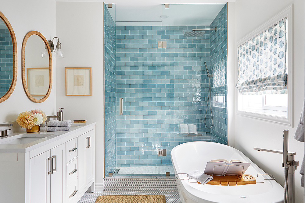 A spacious bathroom with a spa-like feel, thanks to its calming baby blue subway tile backsplash, white walls, white cabinets, chrome hardware and wood-tone accents such as mirror frames, photo frames, a wood bathtub tray and a jute rug.