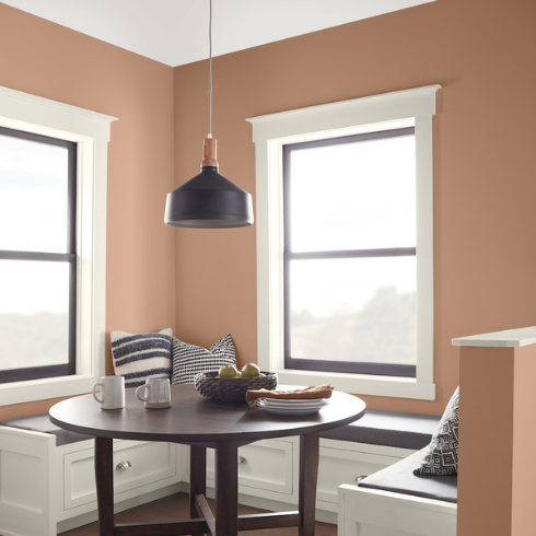 A cozy breakfast nook with a white bench seat covered in grey and white cushion wrapping around a round table send with a fruit bowl, mugs and plates, two big windows, a black pendant lamp, and walls painted with Behr’s Colour of the Year 2021, BEHR Canyon Dusk S210-4