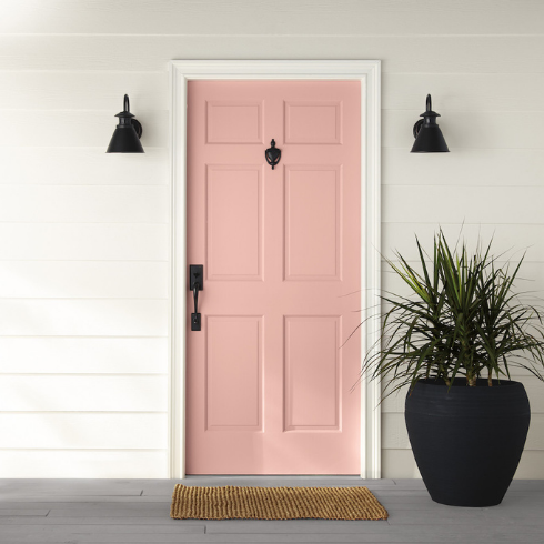 Pink front door painted with BEHR Bubble Shell S160-3 with a black handle and door knocker, two black scones on either side, a large black pot with a palm plant, a coir mat in front of the door, and an exterior wall painted in BEHR Painter's White PPU18-08 and ULTRA PURE WHITE® PPU18-06. IMAGE SOURCE: Instagram: Behr: Bubble Shell S160-3; Painter's White PPU18-08; ULTRA PURE WHITE®