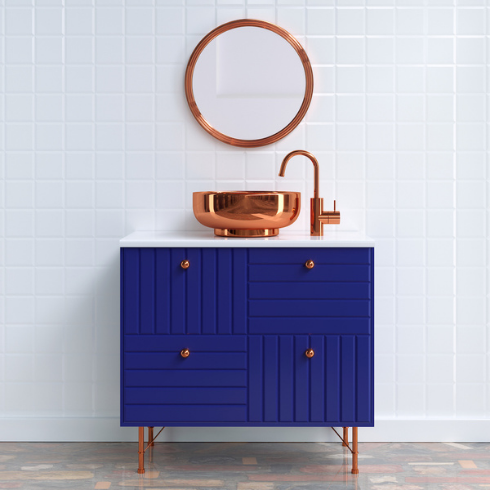 Cobalt blue bathroom vanity with a copper vessel sink and round curved faucet standing in front of a white square tile wall with a round copper framed mirror