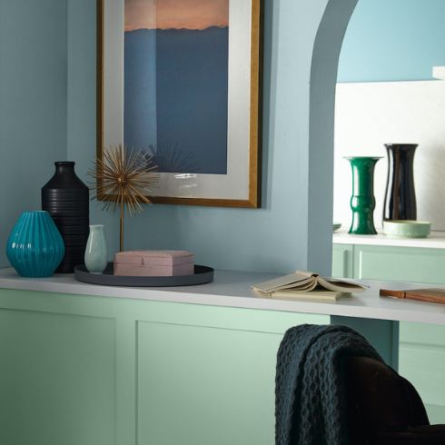 Built-in desk area painted mint green with BEHR Wishful Green M410-2 and decorated with multicolour vases, objets d’arts, a black tray, a pink box, and books and sitting in front of a blue wall painted in BEHR Dayflower MQ3-54 with a gold frame painting hanging on it
