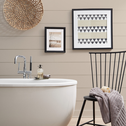 Neutral bathroom with beige-ish walls (BEHR's Almond Whisp), a white standalone bathtub with chrome hardware, a black chair with a towel and luffa, and two black frames and a decorative basket hang on a wood paneled wall painted in BEHR Almond Wisp PPU5-12