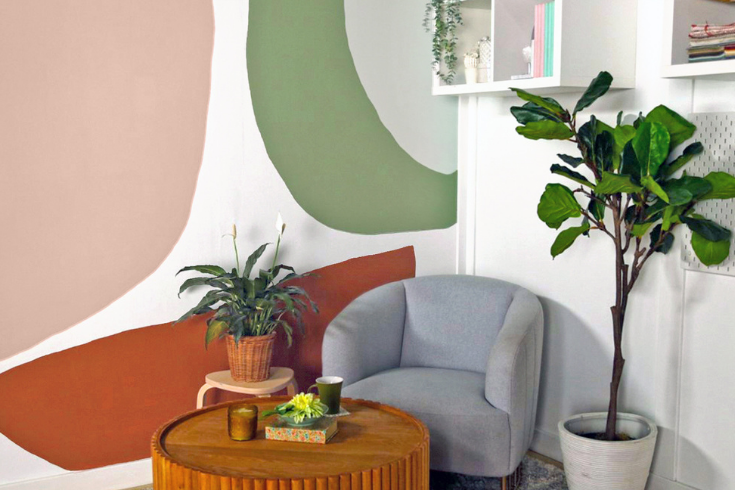 The finished living room with one white accent wall painted all over with large, rounded abstract patterns in pastel colours like pink and sage green. The living room here is still outfitted in the same furnishings as above: A plush grey armchair, indoor plants, and a round wooden coffee table.
