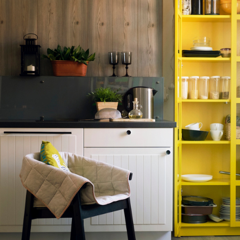 Small modern kitchen with a wood plank backsplash, white shaker lower cabinets with black pulls, black countertops, a metallic industrial lamp hanging from the ceiling, a black painted chair, and a bright yellow crockery cabinet with the door open