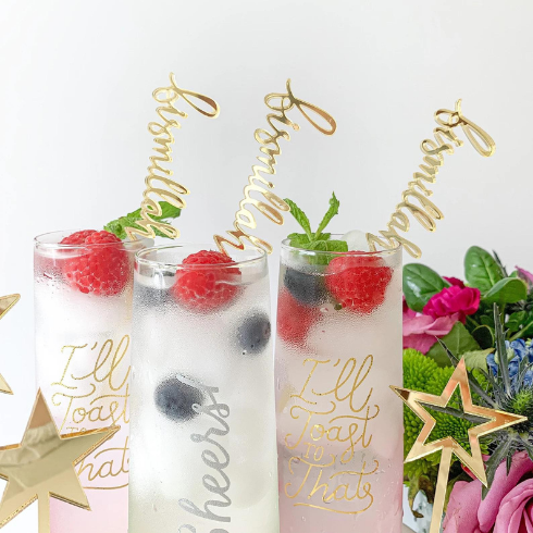 Three tall glasses of iced beverages with raspberries and blueberries, topped with gold stirrers in the shape of the word 