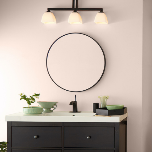 Modern bathroom with a black vanity, black fixtures such as the faucet and overhead light, green and black trays and vases and circular black frame mirror hanging on a light pink wall painted in BEHR Seaside Villa S190-1