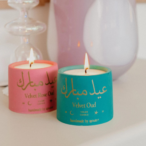 A teal candle and a pink candle side by side, with golden Arabic calligraphy on the votives.