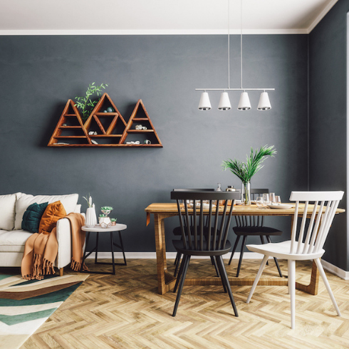 Scandinavian style living and dining room with dark grey painted walls, a three mountain bookshelf on the wall, wooden parquet flooring, a wood dining table with three black and one white chairs around it and white pendant light above it, a white couch with colourful cushions and multicolour rug underneath it