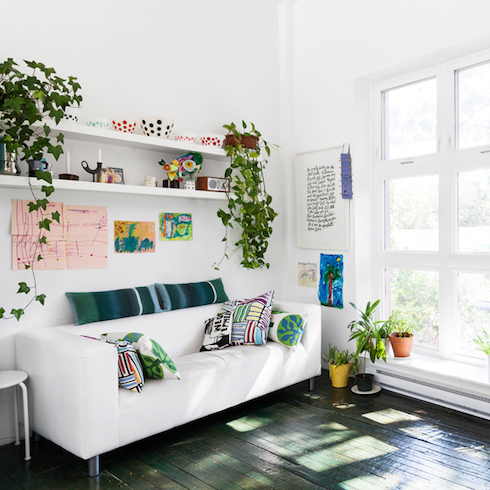 A bright white living room with a large window with plants along the low window sill, a white sofa flanked by more plants, and wall-mounted floating shelves housing even more potted plants.