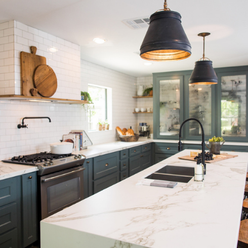 An open kitchen with a large center kitchen island, granite countertops, white subway tile backsplash, two large black pendant lights, and cyan painted cabinets with gold hardware.