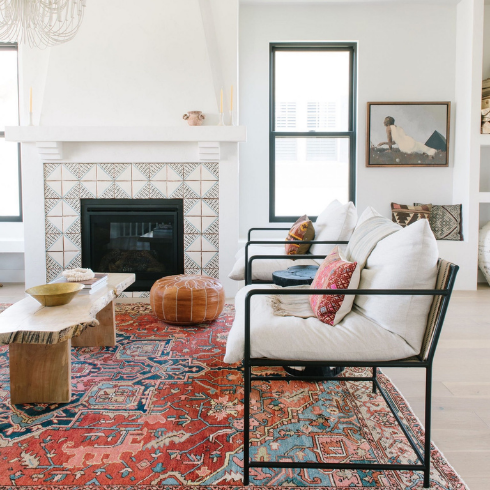 A white living room with boho touches: A large red and teal Persian rug, a live edge wood bench, a tiled border around the fireplace, and patterned cushions on the seating.