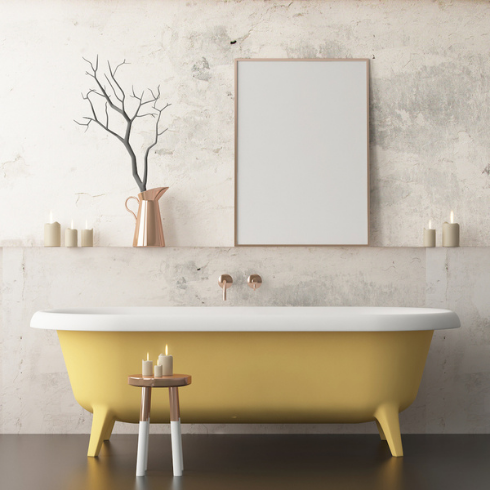 A yellow and white standing bathtub in a modern bathroom with textured marbled wall, copper side table, pitcher vase with a decorative branch, rectangular frame mirror and faucet, and five lit pillar candles.