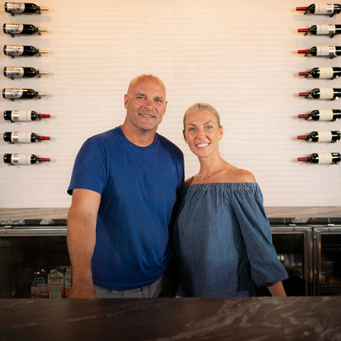 Bryan and Sarah Baeumler standing in front of decorate wall of wine bottles