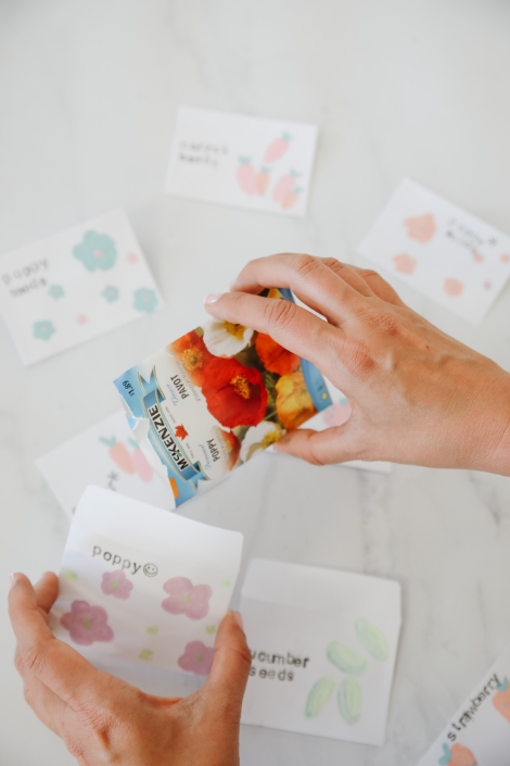 A DIY seed pack labeled "Poppies" and painted with poppy flowers being filled with poppy seeds.