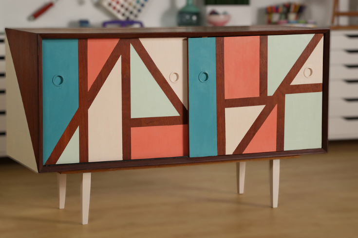 A wooden credenza with geometric patterns painted on its front and sides in pastel colours, and its four wooden legs painted cream.