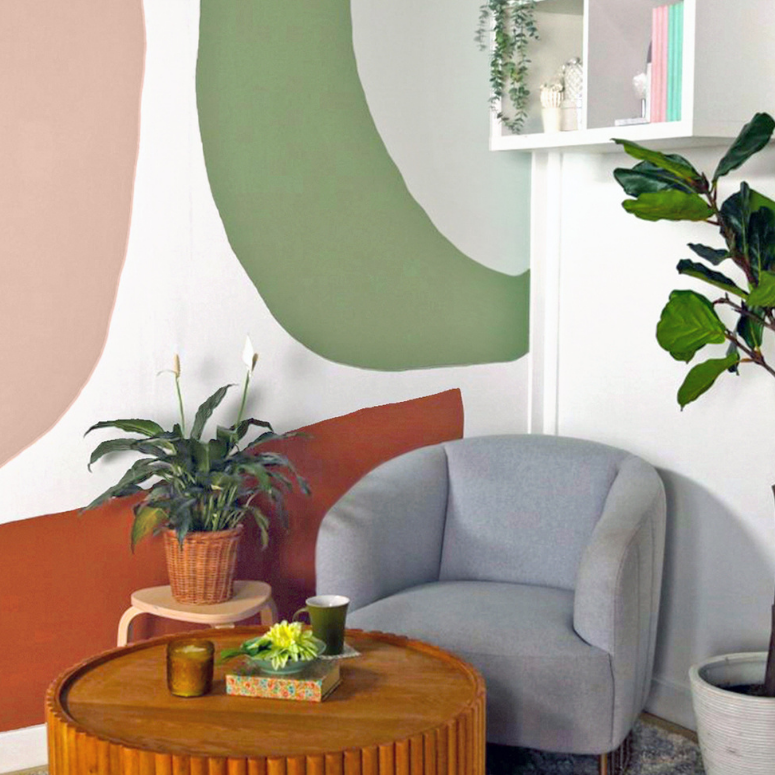 DIY Accent Wall: The finished living room with one white accent wall painted all over with large, rounded abstract patterns in pastel colours like pink and sage green. The living room here is still outfitted in the same furnishings as above: A plush grey armchair, indoor plants, and a round wooden coffee table.