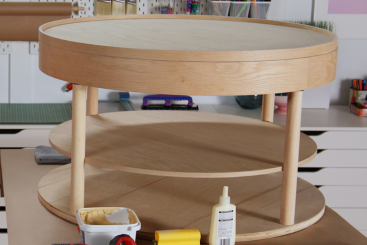A plain wooden round table with three levels, sitting in a workspace with the tools used in this DIY sitting on the floor in front of it.