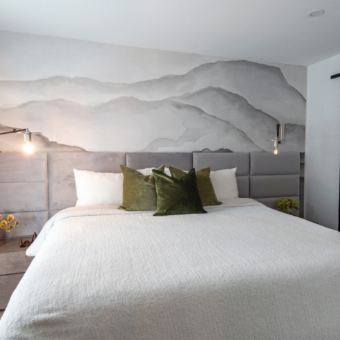 A large bed with white bedding and a wall-mounted grey headboard spanning wall-to-wall. The back wall behind the bed is plastered with a huge grey and white wallpaper mural of mountains.