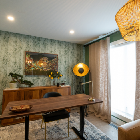 Home office with dark green wallpaper, recessed ceiling lights and a wicker pendant light, dark wood furniture, a tall floor lamp and elegant wall art hanging on the wallpapered wall behind the desk.
