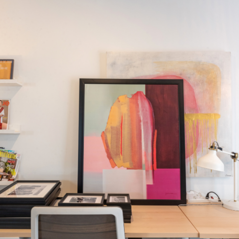 A close-up of Louis' studio desk, with an abstract painting in bold, bright colours like magenta, brown, pale pink, mint green and orange situated on the desk, against the wall.