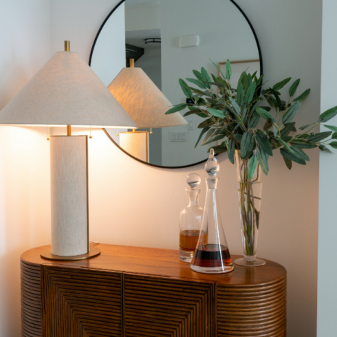 A ribbed wooden credenza. On top of it sits glass decanters of liquor, some greenery cuttings in a tall glass vase and a tall brass and white lamp. A round mirror hangs above.