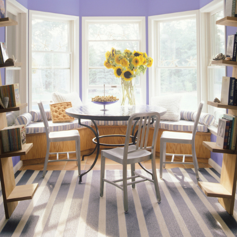 A sunny dining nook with large bow windows with periwinkle-purple walls, built-in wood window benches with upholstery striped in white and shades of purple, a matching rug, a round dining table, white dining chairs, white ladder shelves on either side holding books and knickknacks and a vase of sunflowers on the center of the dining table.