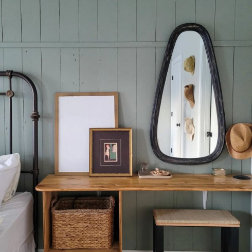 A vertical shiplap bedroom wall painted Sherwin Williams' Evergreen Fog. An oval-ish mirror with a thick black frame hangs on the wall, above a wooden vanity. A black iron bed with white bedding stands beside the vanity, off to one side, partially visible in the picture.
