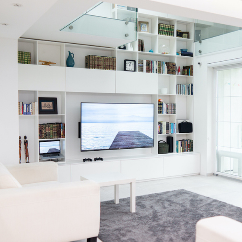 White modern condo living room with a TV and built-in shelves covering a whole wall, a grey rug and white furniture