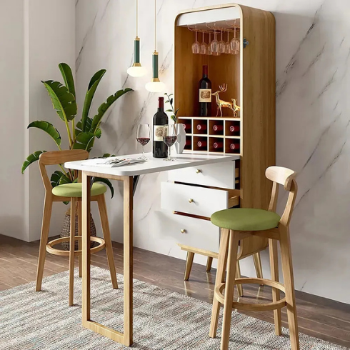 Homary Foldable Bar Cabinet Convertible with Wine Rack & Table & 3 Drawers shown in its open form with countertop, two wood and green stools, a bottle of wine and two wine glasses against a white marble wall with a potted palm tree in the corner